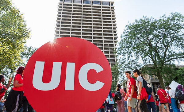 Students walking on campus in front of a large building with a UIC red dot logo in front