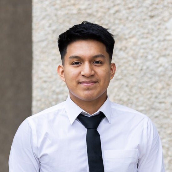 Integrated Health Studies student Paul Lazo in front of tan pebble wall, wearing a white button up with black tie.