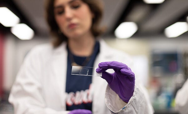 Student holding a glass slide while wearing purple gloves and a lab coat