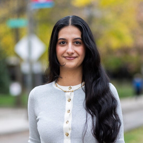 Student with long black wavy hair stands against fall foliage on college campus